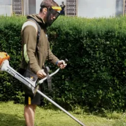 Mężczyzna w masce ochronnej kosi trawę przy pomocy podkaszarki Stihl na tle żywopłotu i budynku z muralem.