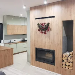 Nowoczesna kuchnia z zabudową w odcieniach beżu i zieleni, wyspą z drewnianym blatem i kominkiem obudowanym drewnem, ozdobionym świątecznym wieńcem.