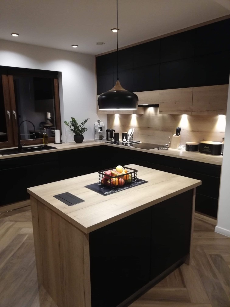 Nowoczesna kuchnia z wyspą, czarne szafki górne i drewnopodobne dolne, oświetlenie punktowe i wisząca lampa nad wyspą z koszykiem owoców