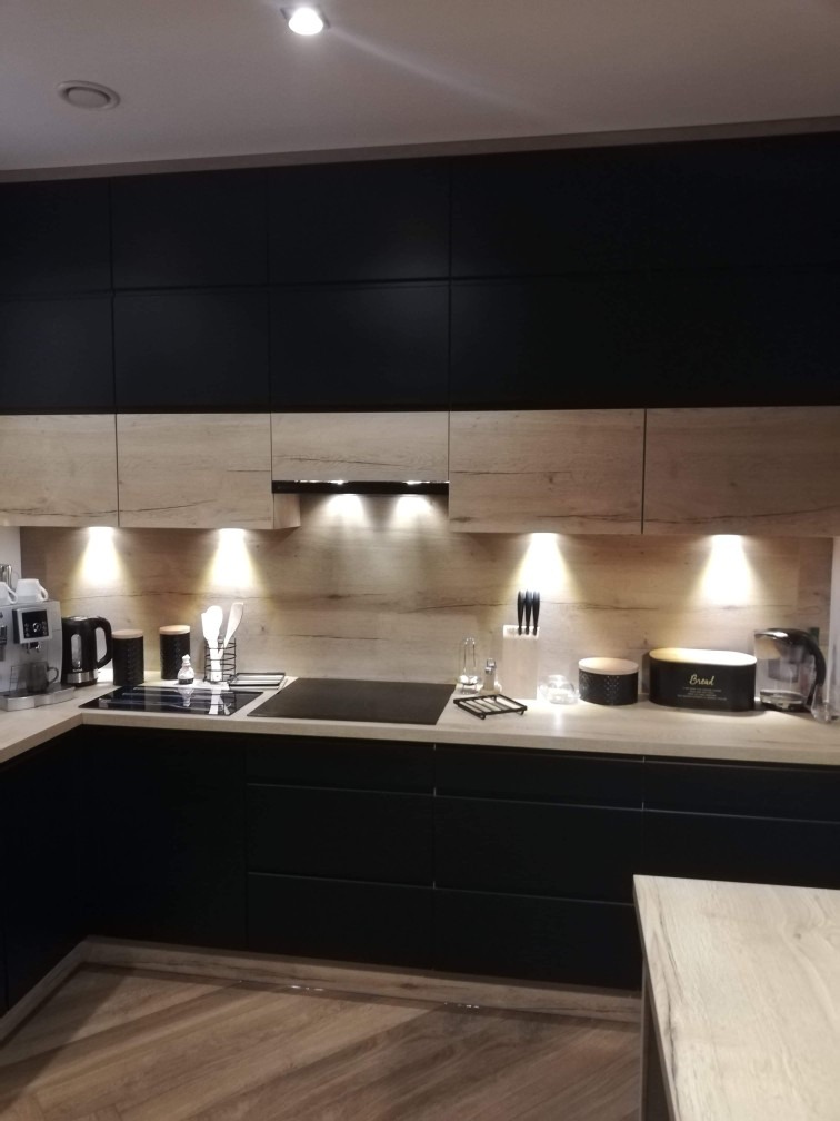 Nowoczesna kuchnia z czarnymi dolnymi szafkami i górnymi szafkami w kolorze jasnego drewna, oświetlona punktowymi lampami podszafkowymi, z widocznym sprzętem AGD i akcesoriami kuchennymi na blacie.