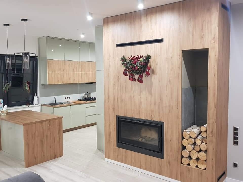 Nowoczesna kuchnia z zabudową w odcieniach beżu i zieleni, wyspą z drewnianym blatem i kominkiem obudowanym drewnem, ozdobionym świątecznym wieńcem.