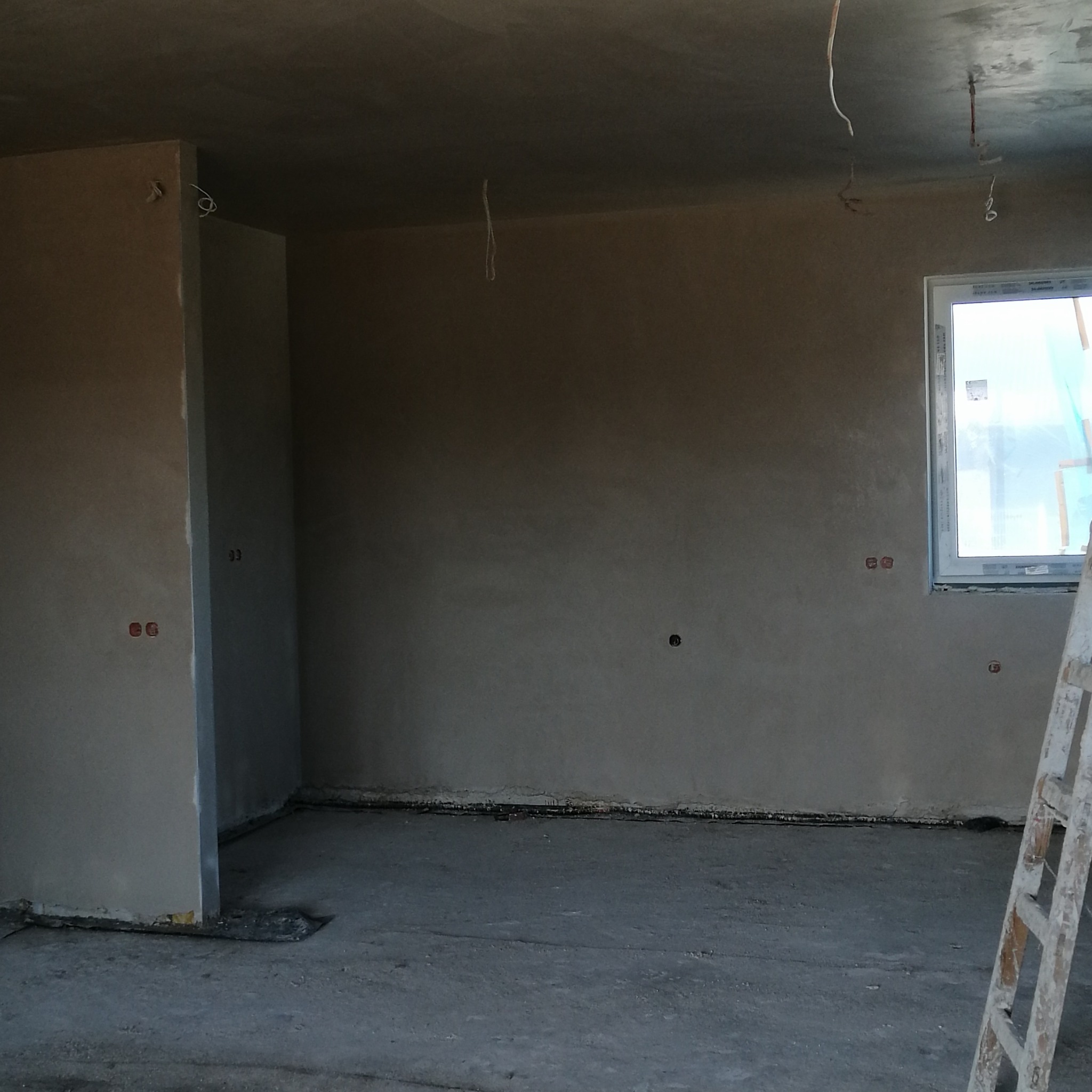 Wnętrze pomieszczenia w trakcie prac wykończeniowych, ściany pokryte świeżym tynkiem, widoczne wystające przewody elektryczne i drabina oparta o ścianę przy oknie.