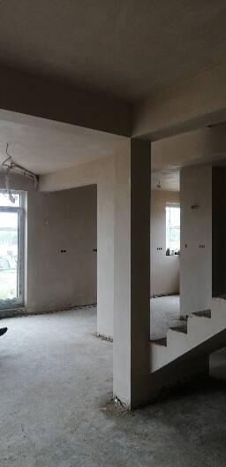 Wnętrze w trakcie prac wykończeniowych: surowe ściany pokryte świeżym tynkiem, betonowa podłoga, widoczne okna i filary konstrukcyjne, schody w stanie surowym, brak umeblowania.