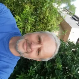 Mężczyzna w niebieskiej koszulce z napisem 'bronzo' na tle zielonej roślinności i fragmentu budynku. Zdjęcie obrócone.