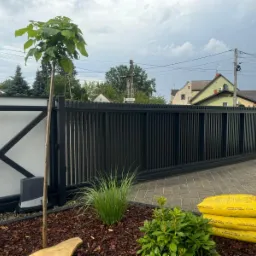 Czarna, przesuwna brama z pionowych paneli, z automatyką, na tle białego muru z czarnym wzorem X, obok świeżo posadzony katalpa i worki kory sosnowej.