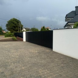 WARMAX GROUP SPÓŁKA Z OGRANICZONĄ ODPOWIEDZIALNOŚCIĄ - Nowoczesne, czarne, pionowe panele ogrodzeniowe zamontowane pomiędzy białym murem, widok z kostki brukowej na posesję w pochmurny dzień.