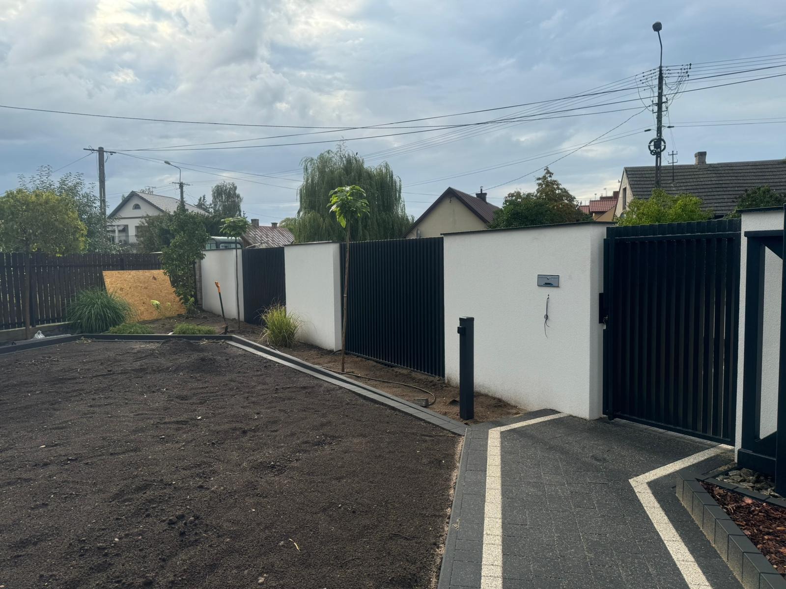 Nowoczesne ogrodzenie z paneli w kolorze antracytowym, połączone z białym murem, widok z przygotowanego pod trawnik terenu, z fragmentem wybrukowanej ścieżki.