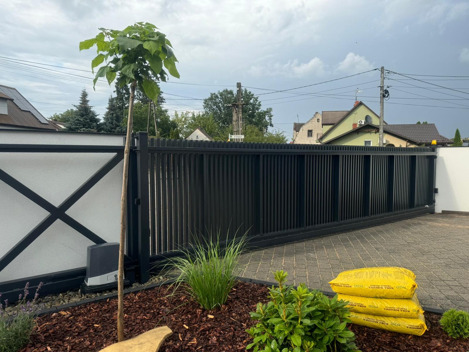 Czarna, przesuwna brama z pionowych paneli, z automatyką, na tle białego muru z czarnym wzorem X, obok świeżo posadzony katalpa i worki kory sosnowej.