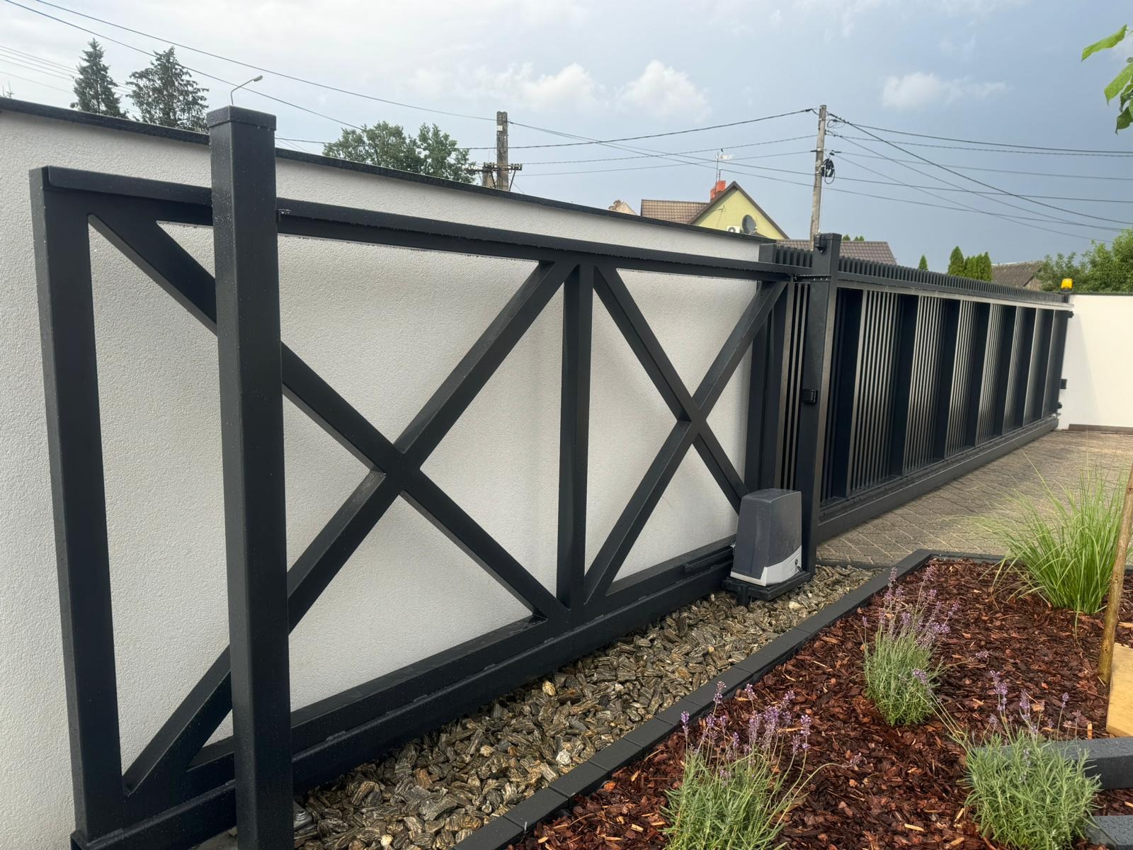 Czarna, przesuwna brama metalowa z dekoracyjnym wzorem krzyży na białym murze, widoczny napęd automatyczny i ozdobny ogródek z lawendą.