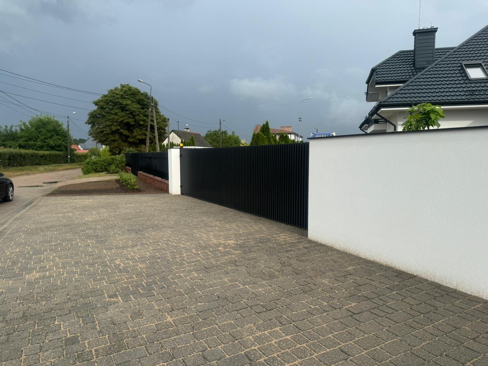 Nowoczesne, czarne, pionowe panele ogrodzeniowe zamontowane pomiędzy białym murem, widok z kostki brukowej na posesję w pochmurny dzień.