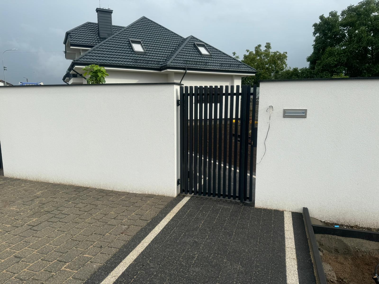 Nowoczesna, czarna furtka z pionowymi szczeblami, wbudowana w biały, otynkowany mur ogrodzeniowy z zamontowaną skrzynką na listy, na tle domu z ciemną dachówką.