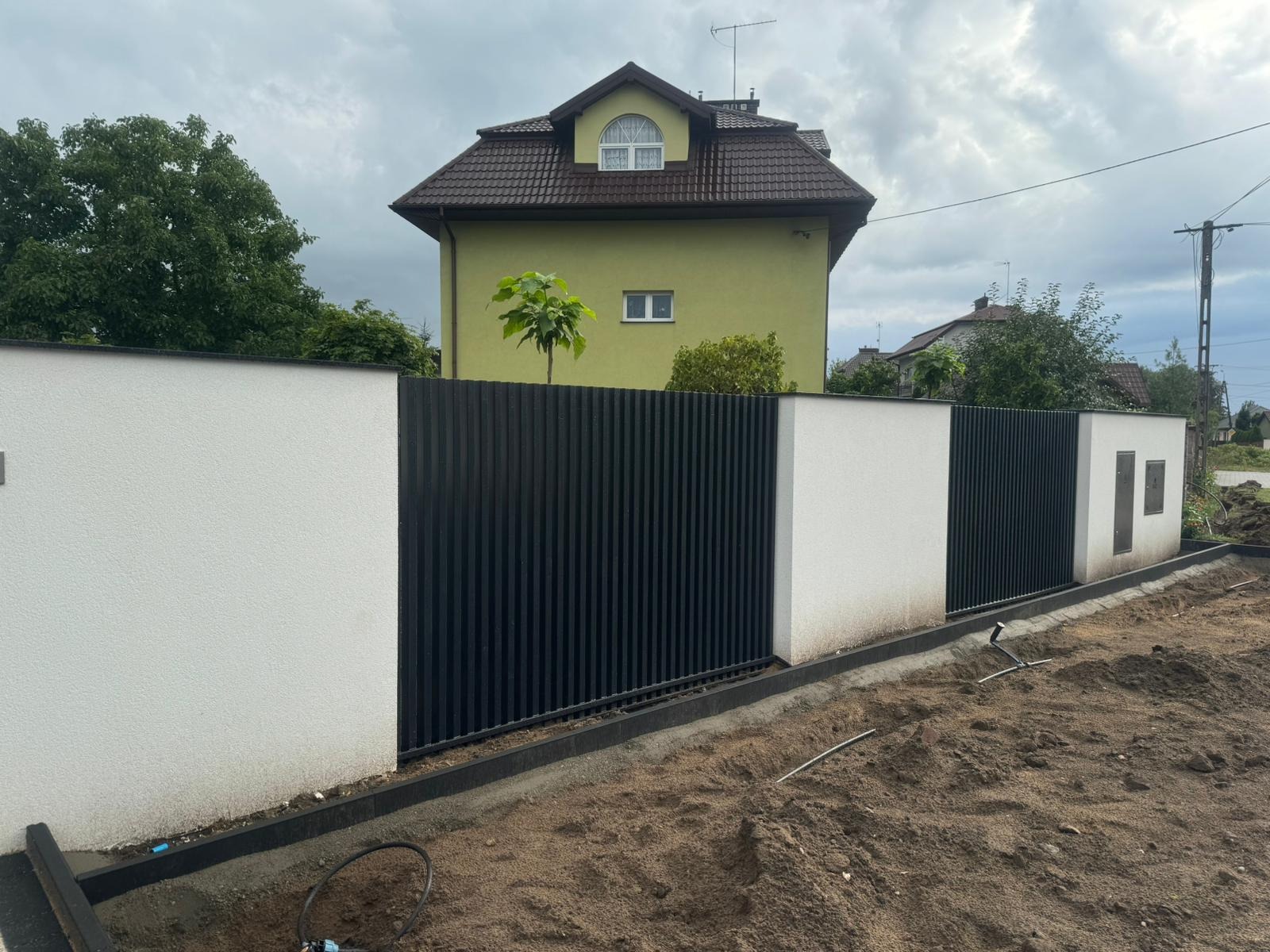 Nowoczesne ogrodzenie z czarnych paneli pionowych i białych słupków, świeżo zamontowane, widoczny teren wokół w trakcie prac ziemnych.