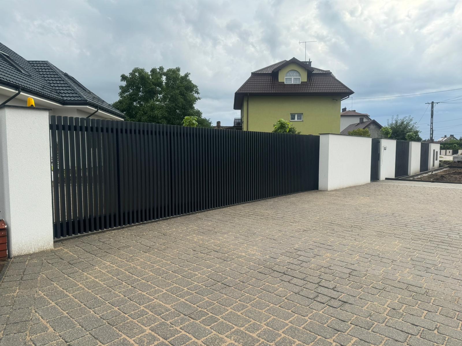 Nowoczesne, czarne ogrodzenie z pionowych profili między białymi słupkami na posesji z kostki brukowej, w tle domy i pochmurne niebo.