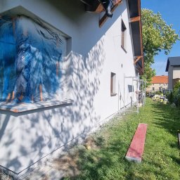 KAROL POGONOWSKI POGON 2 - Boczny widok otynkowanego domu w słoneczny dzień; okno zabezpieczone folią, widoczne rusztowanie i trawnik. Świeżo otynkowana elewacja domu.