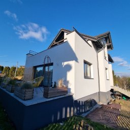 KAROL POGONOWSKI POGON 2 - Biała elewacja domu jednorodzinnego z ciemnymi oknami, widok z boku, w tle błękitne niebo. Widoczne drewniane donice z roślinami na tarasie.