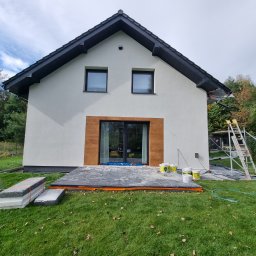 KAROL POGONOWSKI POGON 2 - Ocieplanie domu: biała elewacja, czarna stolarka, przygotowane styropiany, farby i rusztowania na trawie. Prace w toku, widoczne zabezpieczenia.