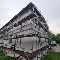 KAROL POGONOWSKI POGON 2 - Dom w trakcie prac elewacyjnych, otoczony rusztowaniem. Widoczne fragmenty elewacji w jasnym odcieniu, samochody w tle i fragmenty zieleni. Ujęcie z dołu.