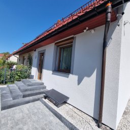 KAROL POGONOWSKI POGON 2 - Biała elewacja domu jednorodzinnego z brązowymi rynnami i schodami z kostki brukowej. Widoczne okno z roletą i elementy systemu alarmowego.