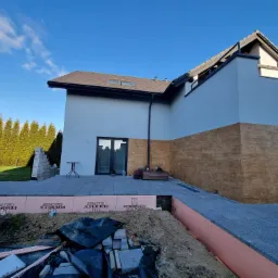 Elewacja domu z fragmentem ocieplenia i wykończeniem imitującym drewno. Widoczny taras z kostki brukowej i materiały budowlane. Częściowo pochmurne niebo.