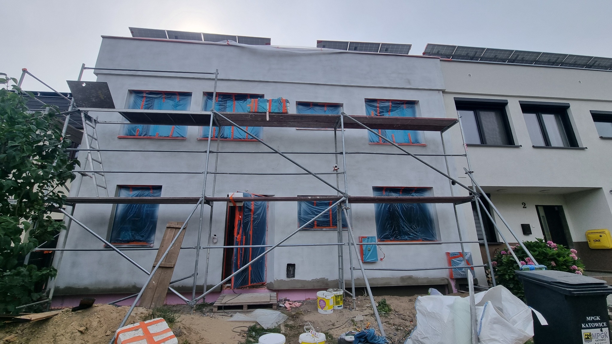 Dom w trakcie tynkowania: rusztowania, okna zabezpieczone folią, świeżo otynkowana ściana. Widoczne panele solarne na dachu i worki z materiałami budowlanymi.
