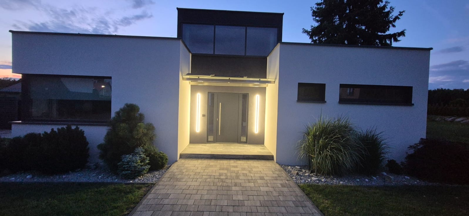 Nowoczesny dom jednorodzinny z oświetloną elewacją i brukowaną ścieżką wejściową o zmierzchu. Minimalistyczny design, szara stolarka, równomierne oświetlenie elewacji.