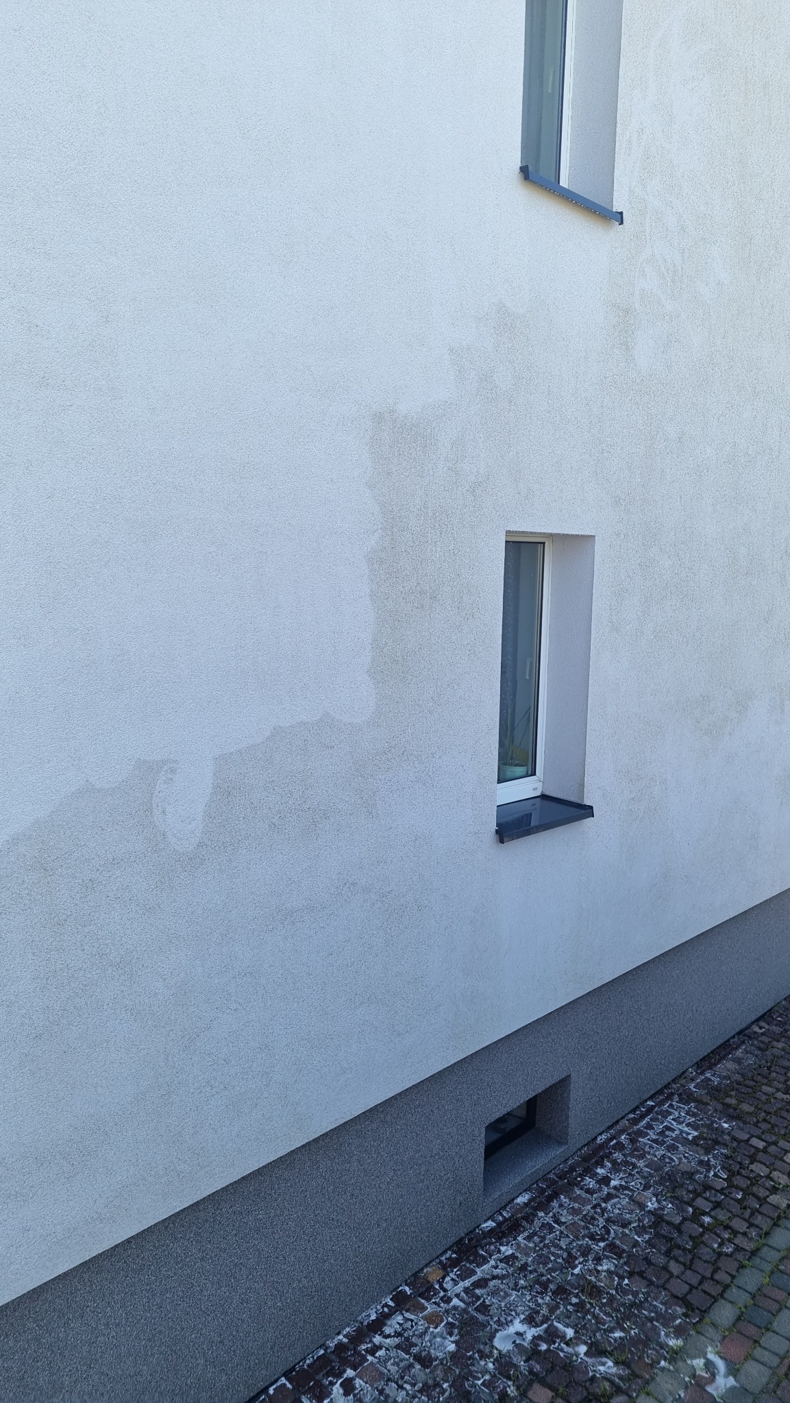 Boczny widok elewacji budynku z widocznymi zaciekami i zabrudzeniami, ukazujący potrzebę mycia elewacji. Fragment okna i kostki brukowej.