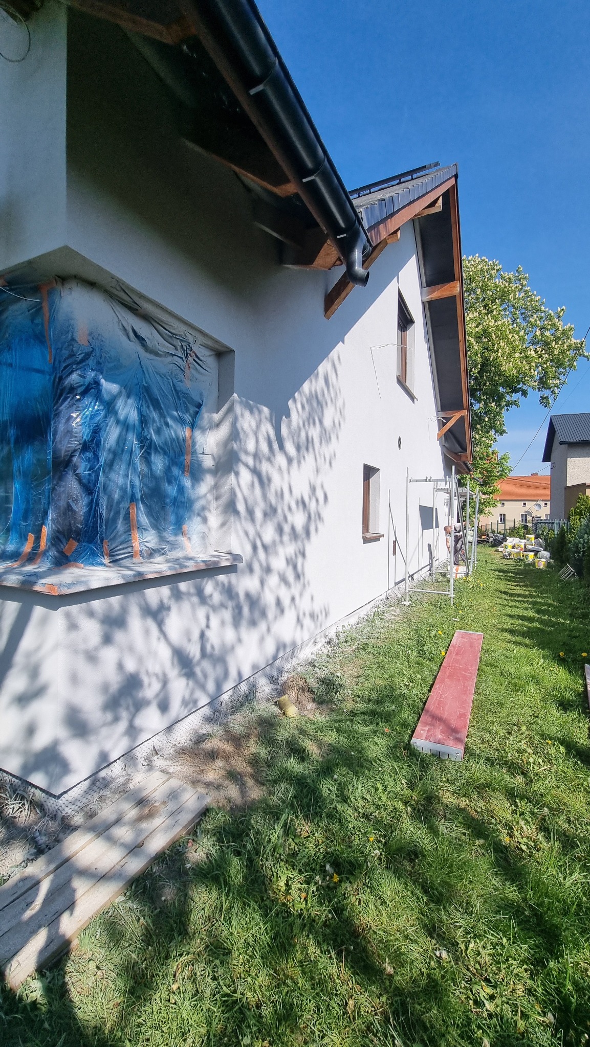 Boczny widok otynkowanego domu w słoneczny dzień; okno zabezpieczone folią, widoczne rusztowanie i trawnik. Świeżo otynkowana elewacja domu.