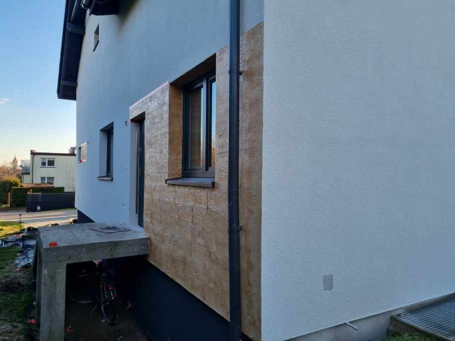 Fragment elewacji domu z dekoracyjnym tynkiem imitującym drewno, widoczny z boku. Część ściany pomalowana na biało, rynna pionowa. W tle budynek i zieleń.