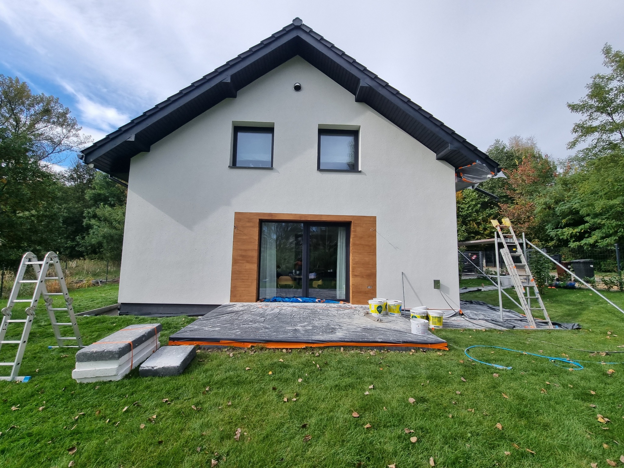 Ocieplanie domu: biała elewacja, czarna stolarka, przygotowane styropiany, farby i rusztowania na trawie. Prace w toku, widoczne zabezpieczenia.