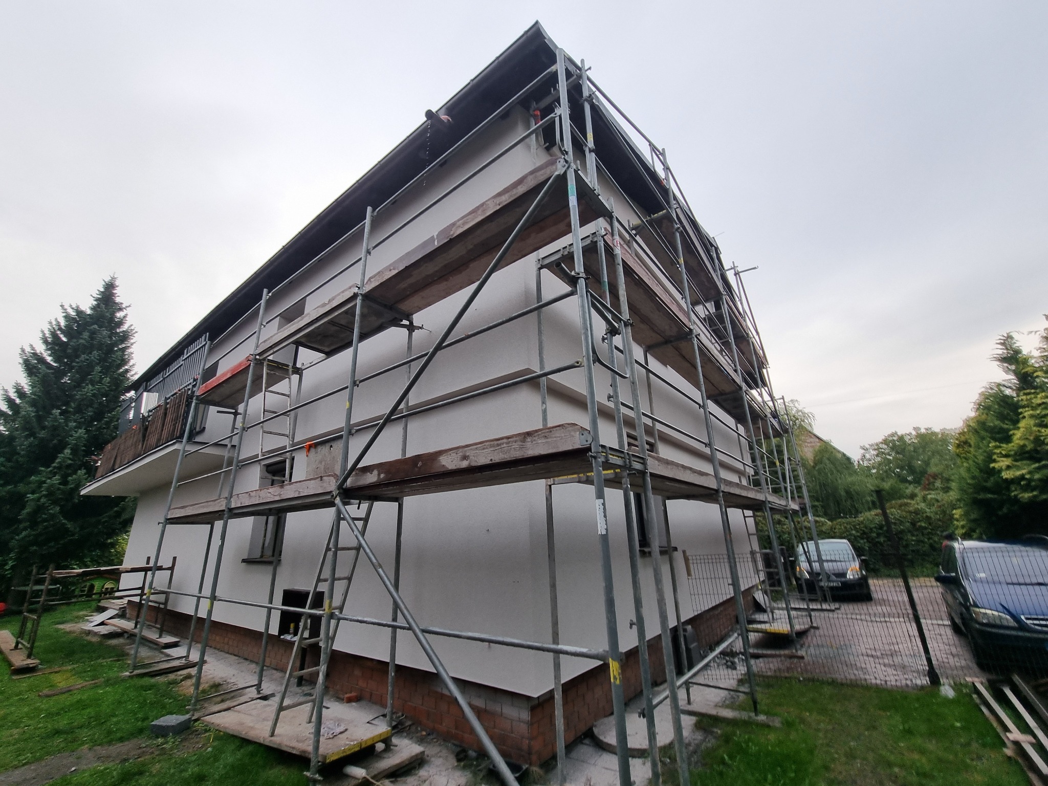 Dom w trakcie prac elewacyjnych, otoczony rusztowaniem. Widoczne fragmenty elewacji w jasnym odcieniu, samochody w tle i fragmenty zieleni. Ujęcie z dołu.