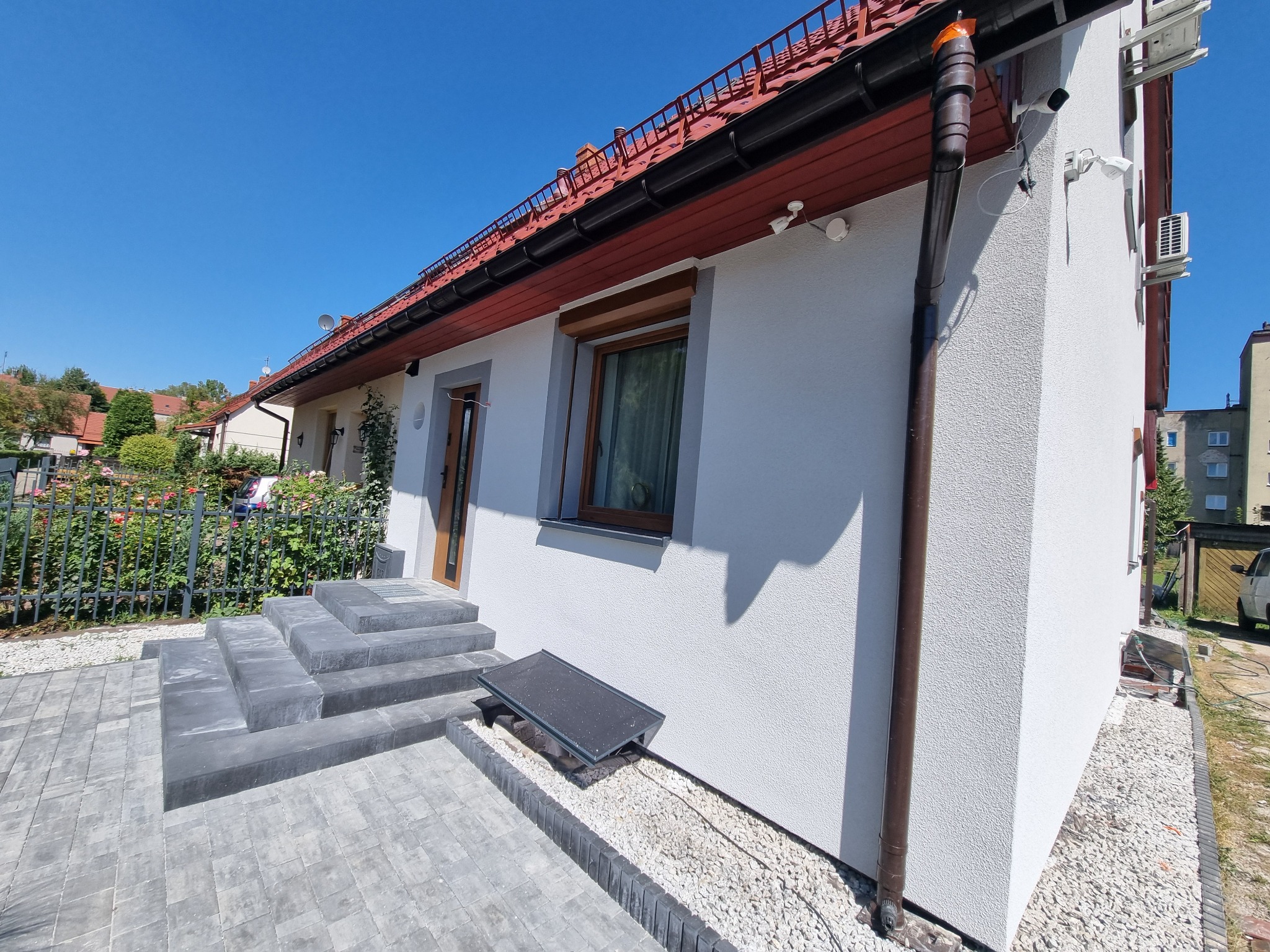 Biała elewacja domu jednorodzinnego z brązowymi rynnami i schodami z kostki brukowej. Widoczne okno z roletą i elementy systemu alarmowego.