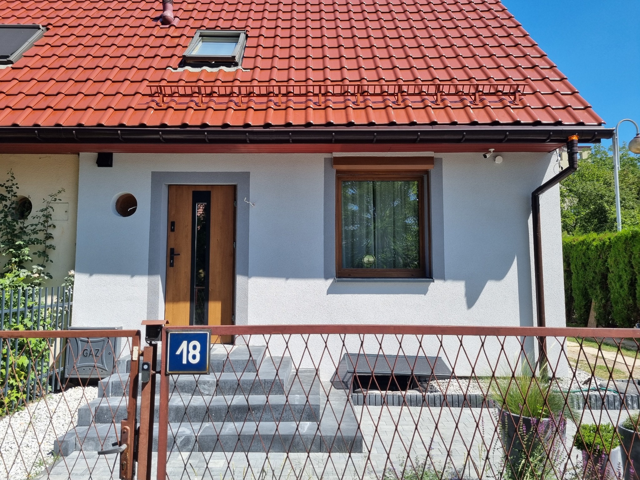 Jasnoszary dom z czerwoną dachówką, brązowymi oknami i drzwiami, numer domu '18' na ogrodzeniu. Widok z przodu, w słoneczny dzień.