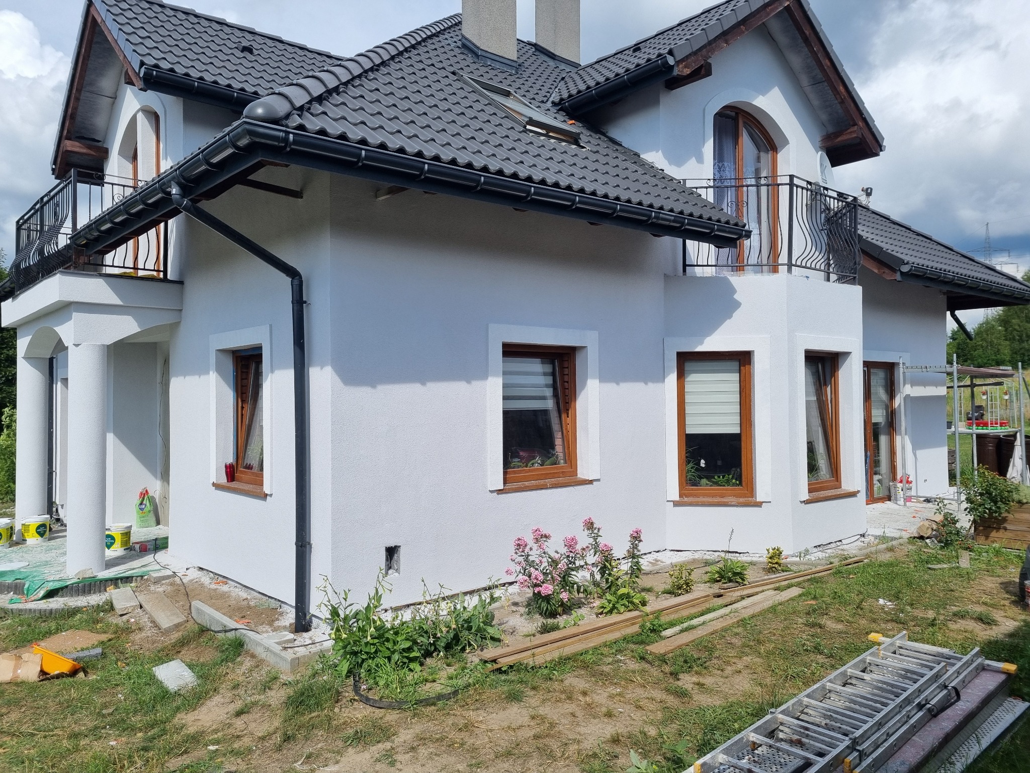 Biały dom z szarym dachem, widok z boku, z oknami z brązowymi ramami. Świeżo otynkowana elewacja, widoczne narzędzia i materiały budowlane.