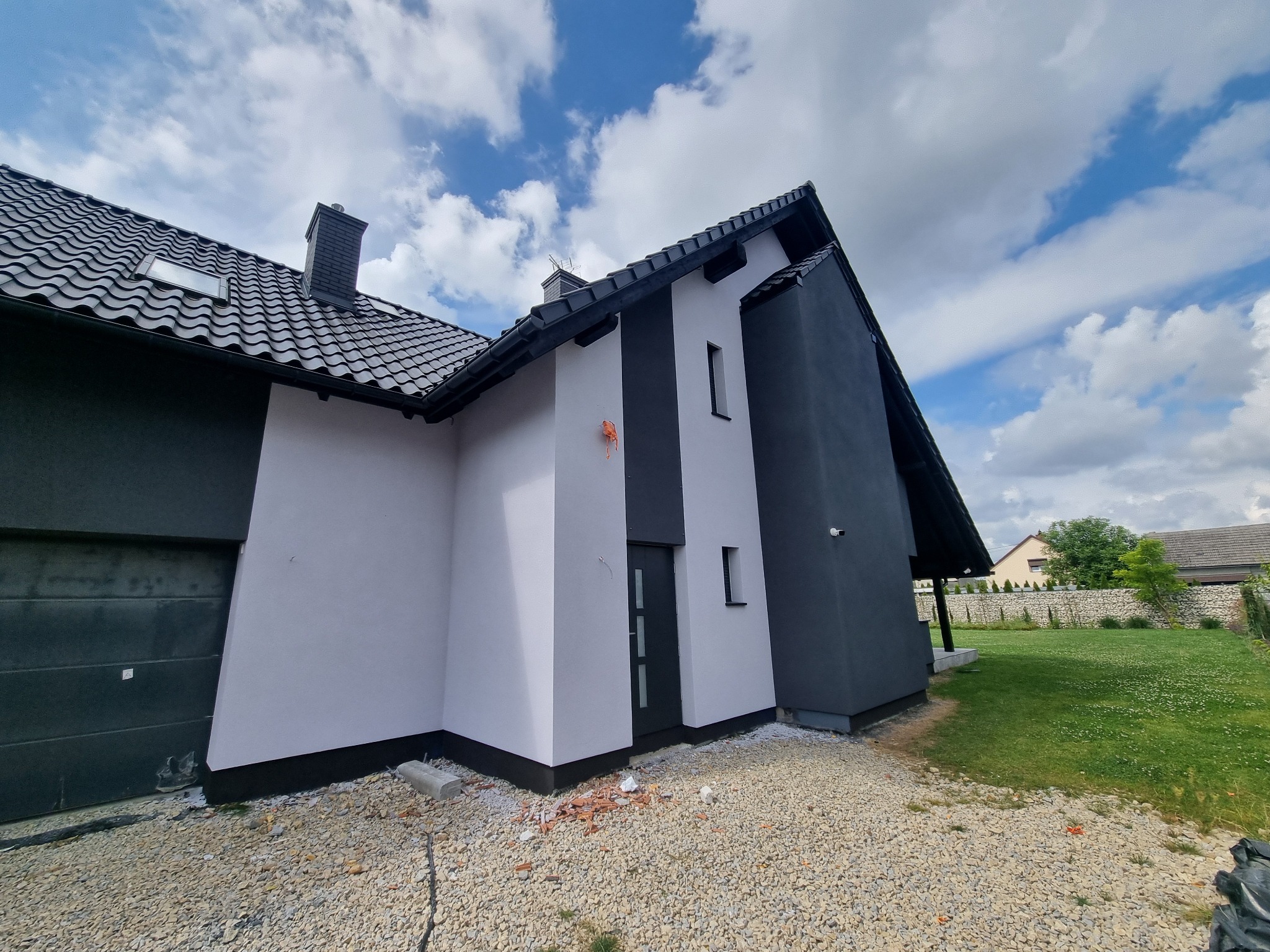 Nowoczesny dom jednorodzinny z dwukolorową elewacją (szary i biały), czarną dachówką i elementami wykończeniowymi. Widoczne detale: okno dachowe i fragment elewacji.