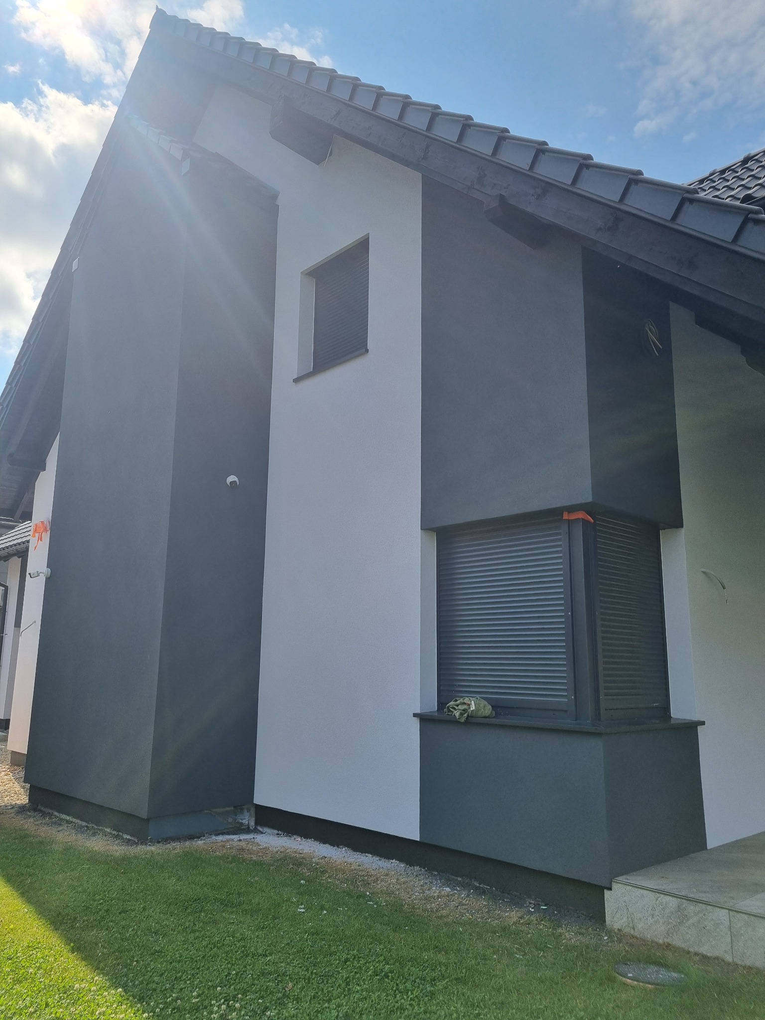 Dwukolorowa elewacja domu z roletami w Będzinie. Szara i biała kolorystyka, widoczne okna z roletami zewnętrznymi, trawnik. Ujęcie z dołu, widać fragment dachu.