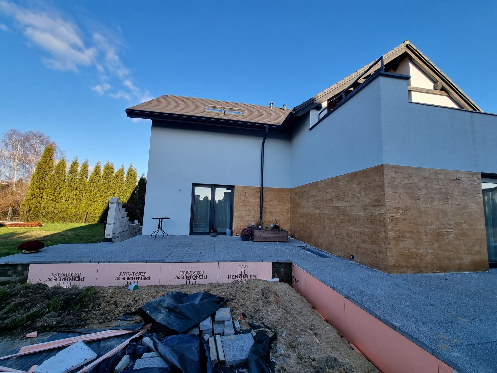 Elewacja domu z fragmentem ocieplenia i wykończeniem imitującym drewno. Widoczny taras z kostki brukowej i materiały budowlane. Częściowo pochmurne niebo.