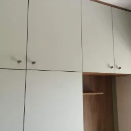 Biała szafa wnękowa z drewnianym wykończeniem g&oacute;rnej krawędzi i wnętrzem, z okrągłymi, metalowymi uchwytami na frontach.