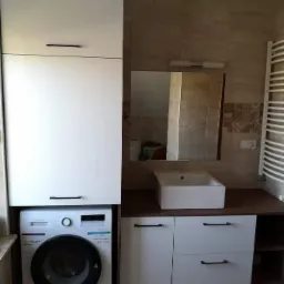 Biała szafka łazienkowa na wymiar z pralką w zabudowie, umywalka nablatowa i płytki imitujące kamień na ścianie.