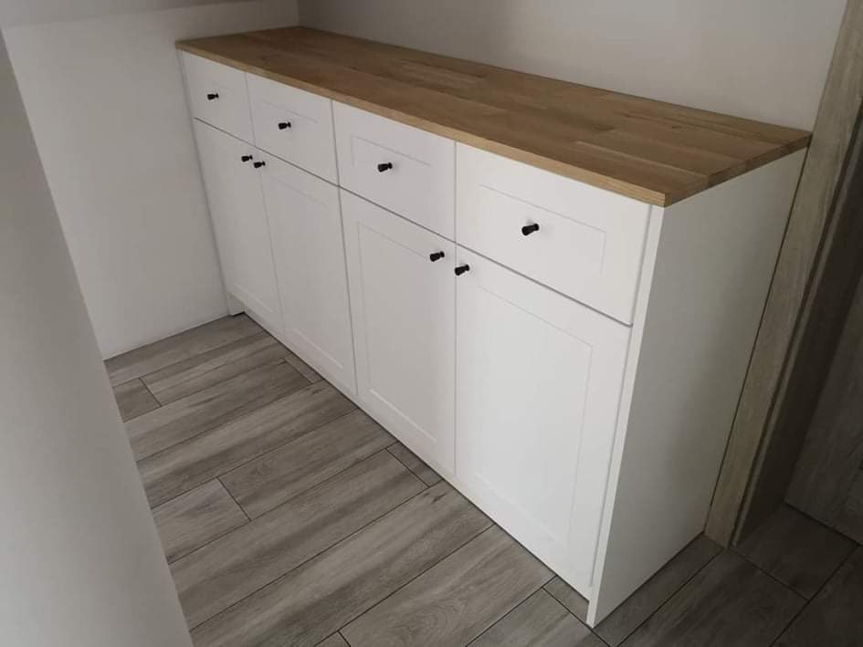 Biała szafa wnękowa z drewnianym blatem, czarne gałki, szare płytki podłogowe imitujące drewno.