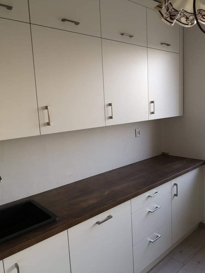 Jasne szafki kuchenne z ciemnym blatem drewnianym i czarnym zlewozmywakiem, widoczny fragment ozdobnej lampy sufitowej.