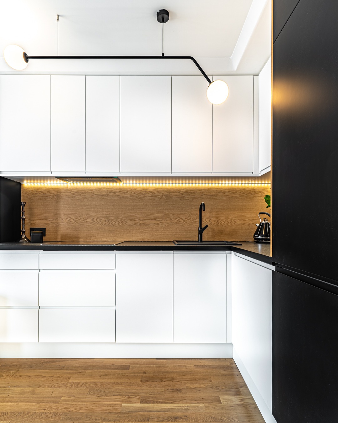 Nowoczesna, minimalistyczna kuchnia w Warszawie z białymi szafkami, czarnym blatem i drewnianym panelem ściennym podświetlonym LED, oświetlona designerską lampą.
