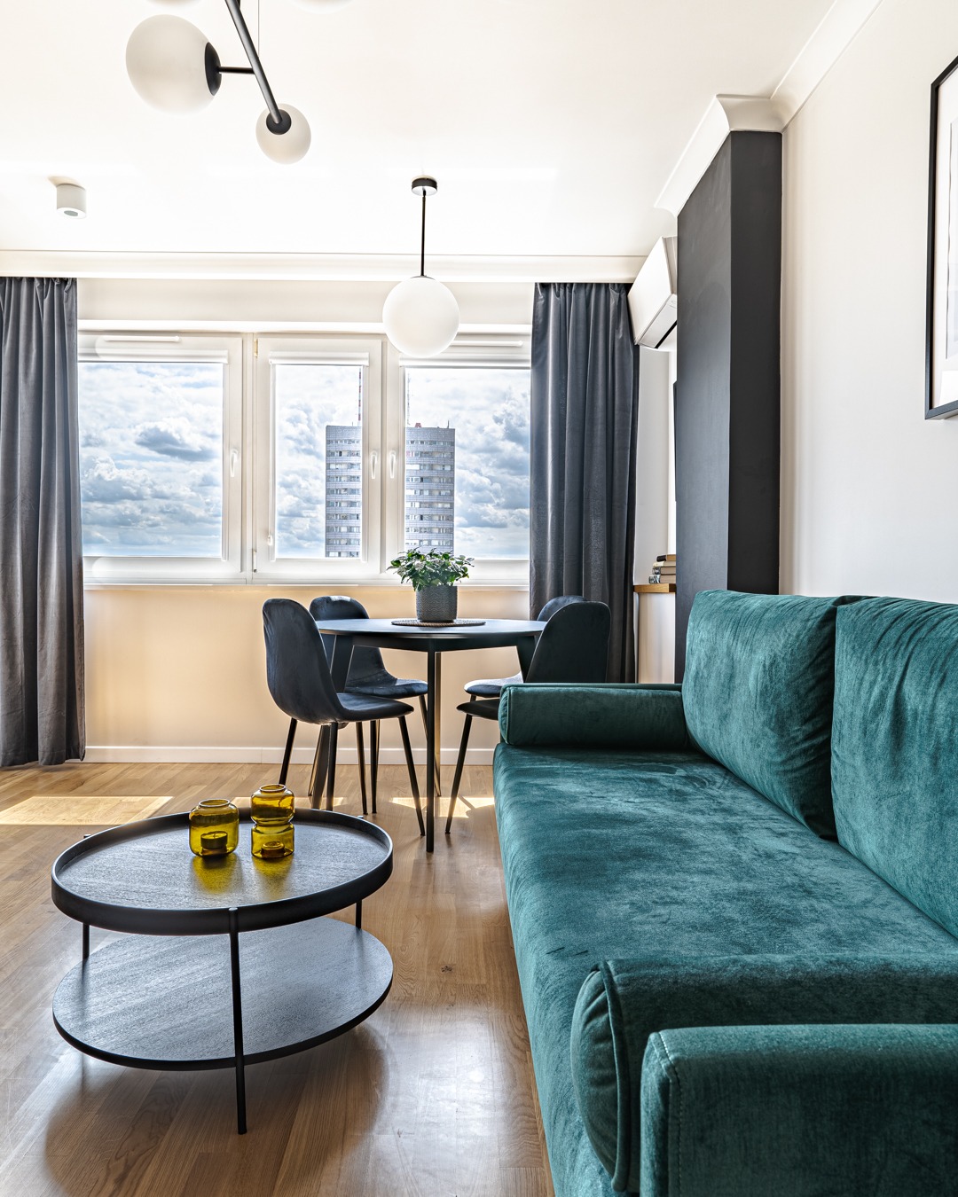 Jasne wnętrze salonu z turkusową sofą, okrągłym stolikiem kawowym z bursztynowymi świecznikami, okrągłym stołem jadalnym z kwiatem i trzema oknami z widokiem na miasto.