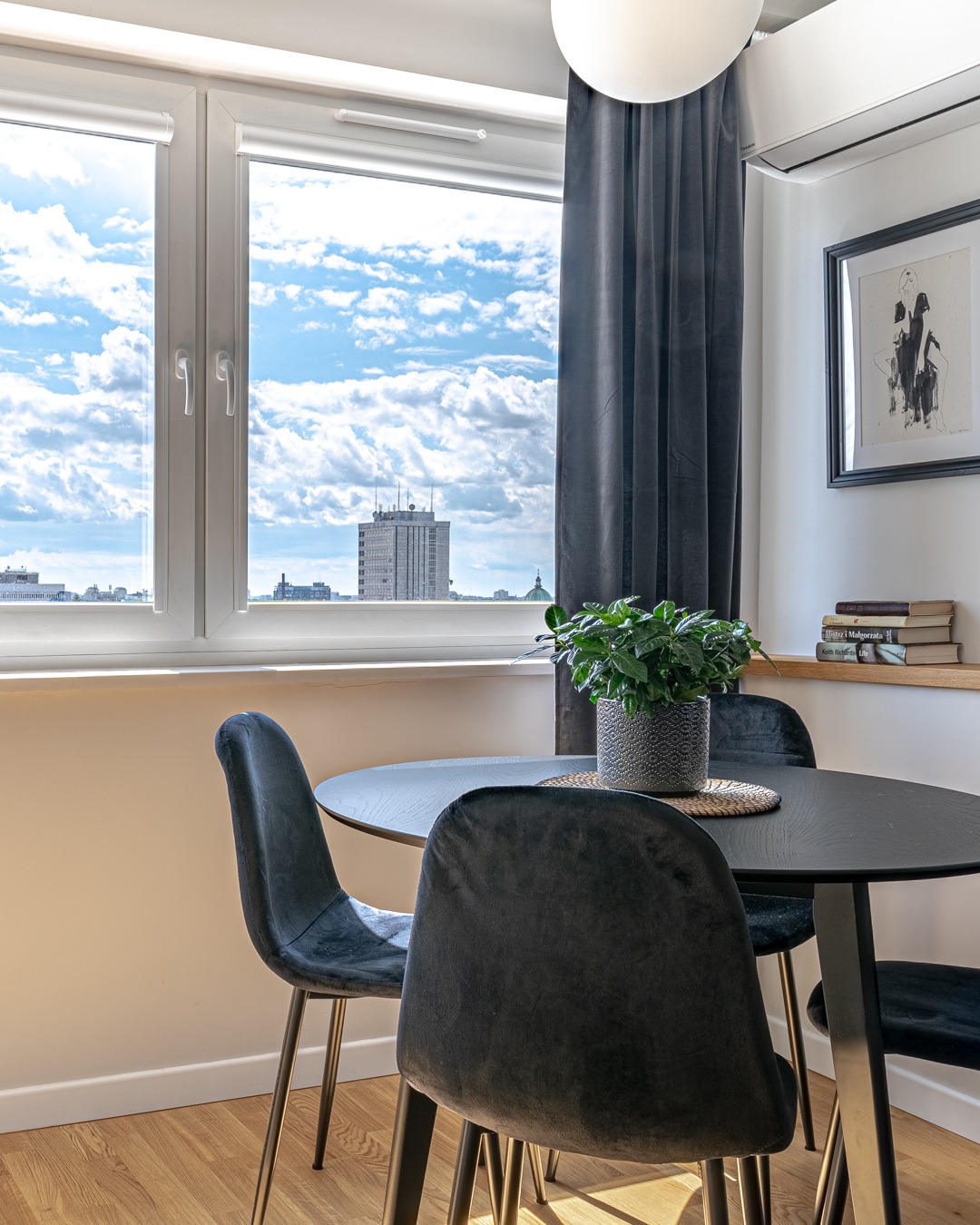 Jadalnia w mieszkaniu: okrągły, czarny stół z welurowymi krzesłami na złotych nóżkach, doniczka z zieloną rośliną na środku, widok na miasto zza okna.