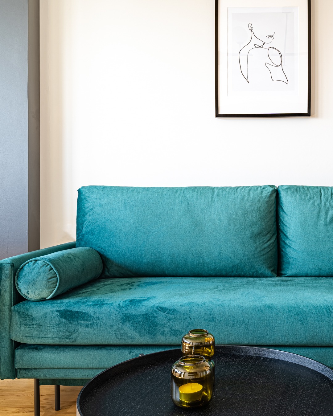 Turkusowa, welurowa sofa z poduszką-wałkiem, czarny stolik kawowy z dwoma świecami w szklanych lampionach, grafika w czarnej ramie na białej ścianie, minimalistyczne wnętrze.