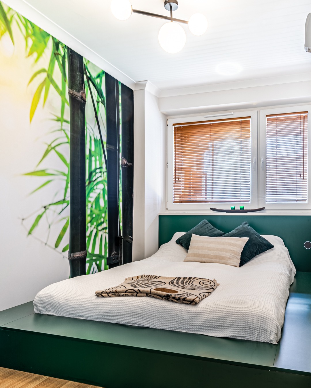 Nowoczesna sypialnia z łóżkiem na podwyższeniu w kolorze butelkowej zieleni, ściana z fototapetą bambusową i drewniane żaluzje w oknach.