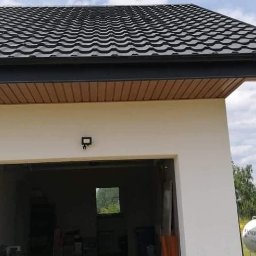 Usługi remontowo-budowlane Łukasz Lamparski - Nowoczesny dom z ciemną dachówką, kremową elewacją, widoczną rynną i zbiornikiem na gaz w tle.