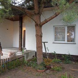 Usługi remontowo-budowlane Łukasz Lamparski - Biała elewacja domu jednorodzinnego z widocznym fragmentem werandy i drzewem iglastym na pierwszym planie, mężczyzna obserwuje postęp prac na werandzie.