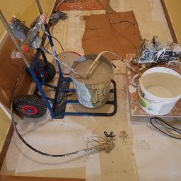 Usługi remontowo-budowlane Łukasz Lamparski - Sprzęt do malowania natryskowego z wiadrem farby na podłodze zabezpieczonej brązowym papierem, widoczne ślady farby na podłodze i urządzeniach.