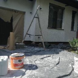Usługi remontowo-budowlane Łukasz Lamparski - Przygotowania do odświeżenia elewacji domu w Kaźmierzynie: widoczna drabina oparta o ścianę, okna zabezpieczone folią, wiadro z farbą i kanister z preparatem na folii ochronnej.