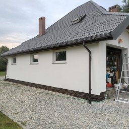 Usługi remontowo-budowlane Łukasz Lamparski - Świeżo pomalowana elewacja domu jednorodzinnego z szarą dachówką, widoczne okna, rynny i stos opon obok, drabina oparta o ścianę garażu.